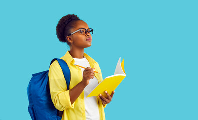 Portrait happy beautiful African American student girl in yellow shirt and eyeglasses standing isolated on blue background, holding pencil and notebook, writing essay, looking up and thinking of ideas