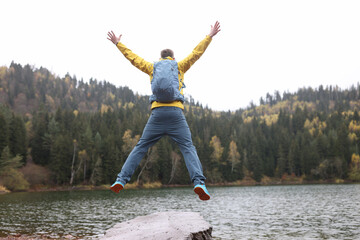 Happy traveler with backpack jumps with open arms, in background lake and mountains.