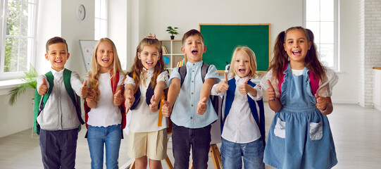 Happy group elementary school students showing thumbs up rejoicing at getting quality education with interesting learning activities stand in spacious classroom. Children and kids back to school
