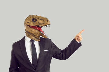 Strange eccentric business man wearing elegant suit and funny ugly masquerade dinosaur monster mask standing isolated on light background pointing index finger aside at something interesting on right