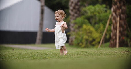 Funny little baby running in the garden on a hot summer day. Cute caucasian baby running on grass in park. Little adorable child happily smiling, expressing emotions - happy childhood concept 