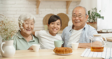 Obraz na płótnie Canvas Mature asian couple and their teen granddaughter sitting together at kitchen table, smiling and looking at camera - family ties concept portrait closeup 