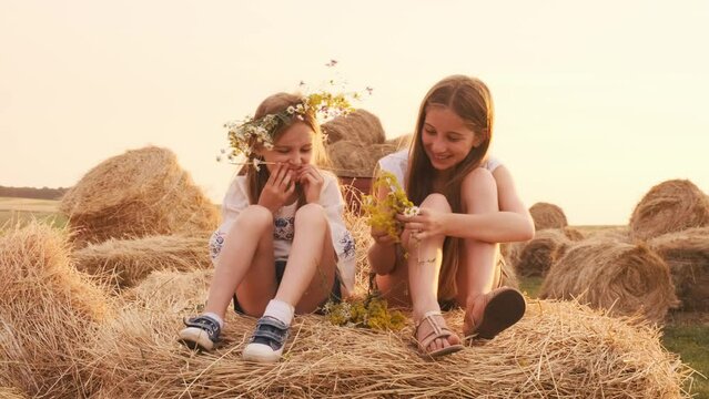 Two pretty sisters wearing wreath sitting on hay stack, having fun and smiling. Girls spending time together in the field in sunset time and making mustache out of flowers.