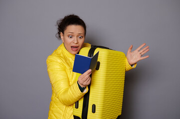 African American woman in bright yellow jacket, carrying yellow suitcase, looking with shock at her passport, expressing disappointment of problems with visa or documents, isolated on gray background - obrazy, fototapety, plakaty