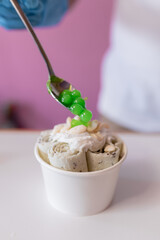 A worker is decorating a rolled ice cream tub with green bubble toppings with a spoon