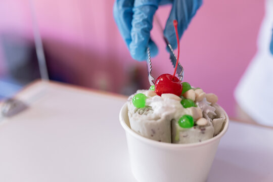 Close up of a worker is decorating a rolled ice cream tub with a cherry on top using tongs