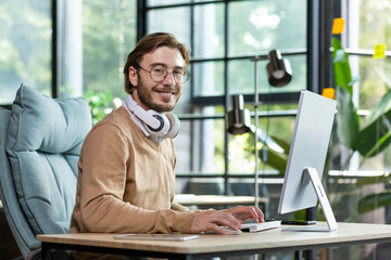 Fototapeta premium Portrait of a young freelancer, programmer sits in the office in white headphones and works at the computer. Smiling looking at the camera.