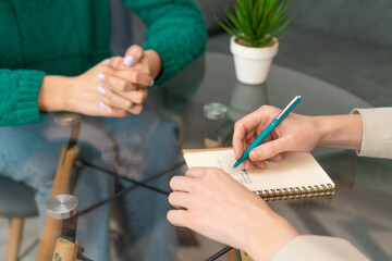 Close-up of a woman's hands with a notebook and a houseplant on the table.Two women are sitting at a glass table, one is taking notes in a notebook. Selective focus