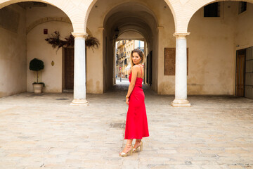 Fototapeta na wymiar Young, blonde, beautiful woman in a red dress is visiting seville. The woman poses for the camera very elegant and like a model in the typical streets of the city. Holidays and travels