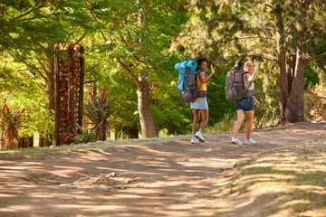 Fototapeta na wymiar Two Female Friends With Backpacks Setting Off On Vacation Hiking Through Countryside Waving