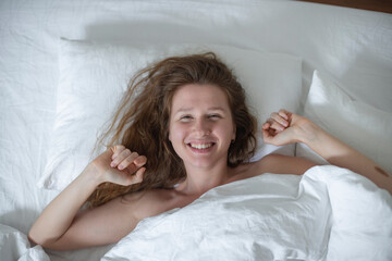 Obraz na płótnie Canvas Young happy beautiful sleepy woman in the bed in bedroom at home in the morning lying under white blanket, enjoy resting, sleep well, wake up and smile. Good morning