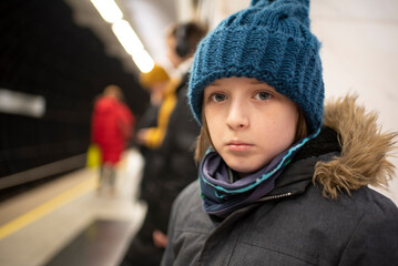 a sad boy in a winter jacket and hat is waiting for an approaching train on a subway platform