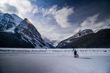 people ice skating on frozen and covered with snow lake luise during winter with mountains on...
