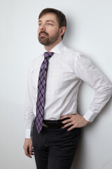 Studio portrait of a middle aged man wearing a white buttoned up dress shirt and a purple pattern tie. He is standing against a wall. He has a beard and brown hair. 