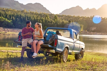 Couple With Backpacks In Pick Up Truck On Road Trip By Lake Drinking Beer