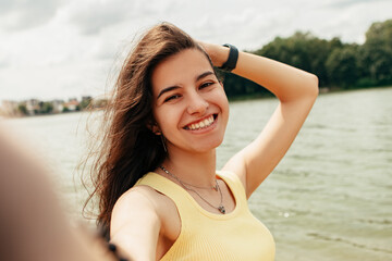 Woman standing smiling laughing near river sea lake, take picture, make selfie hold camera. Female on beach island. Girl tourist on vacation, weekend relaxing resting resort. Lady go shore, sunbathing