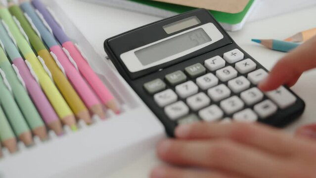 Child hands with calculator counting during school homework and colorful crayons on table. Kid fingers press buttons and calculate doing math closeup