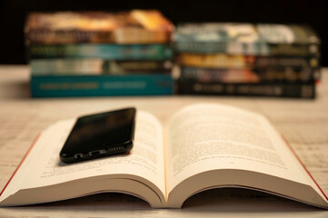 An open book with a smartphone resting on its pages, with blurred stacks of books in the background.