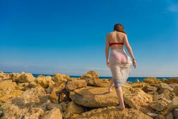 Young woman waving scarf in wind at beach. Happy woman in red bikini holding tissue and looking away at sea. Sexy fashion woman playing with the scarf on rocky seashore.