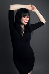 Studio portrait of a caucasian woman in her 30s wearing a black dress and smiling. Her arms are raised over her head in a pose. She has black hair and is wearing red lipstick.