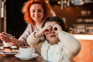Beautiful young red-haired mother with cute curly-haired daughter are drink hot cocoa with cake in a cozy cafe.