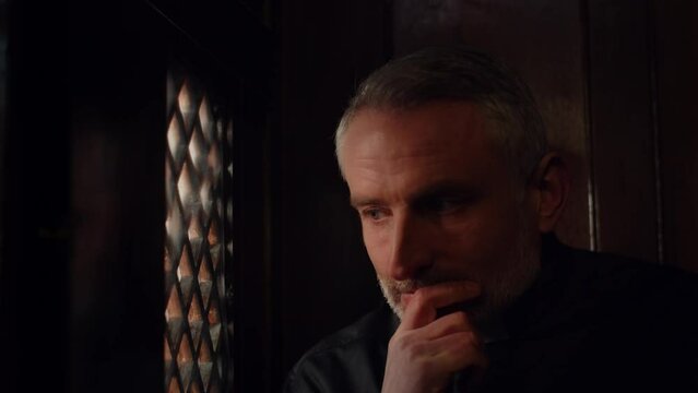 A pastor in a black cassock with a white collar listens attentively to a parishioner. The priest sitting in the confessional listens to the confession.