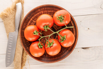 Several fresh tomatoes on branch with metal knife and clay plate on wooden table, macro, top view.