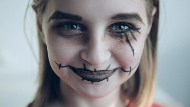 Preteen girl with spooky Halloween makeup opens eyes and smiling looking at camera closeup portrait. Creepy child kid with crazy joker expression acting