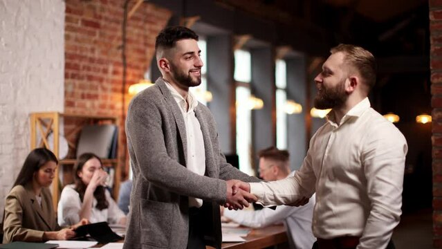 Cooperation and partnership. Two businessmen shaking hands in agreement. Blurred image of employees working on background. Concept of business, career development, brainstorming, teamwork