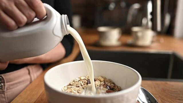 Girl puts milk to oatmeal in bowl plate for delicious cereal breakfast at kitchen. Woman preparing granola for morning healthy diet meal with latte