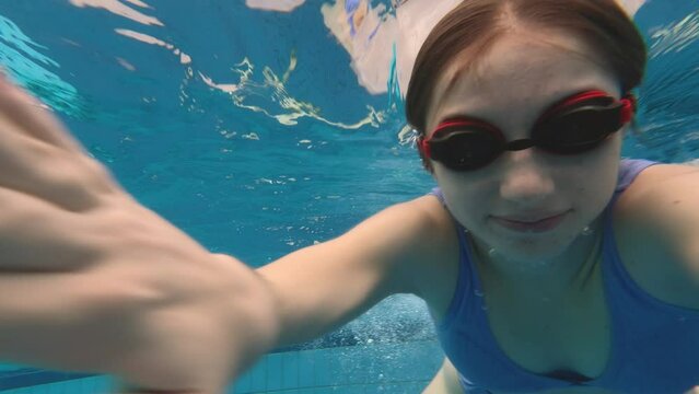 Beautiful girl teenager swimming in pool under water with bubbles wearing glasses googles. Pretty female person with camera diving in blue water and recording herself
