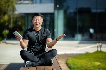 A happy young Asian sportsman man couch is sitting resting on a wooden bench near the lake and using a mobile phone, holding headphones. Smiling at the camera.