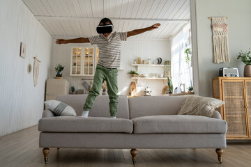 Carefree African boy child using VR helmet for fun and entertainment, happy kid standing on sofa playing immersive 3D virtual flight game, flying in metaverse world. Children in immersive environment