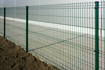 A new green metal wire fence around a new gray industrial facility, soil for grass and a paved lane