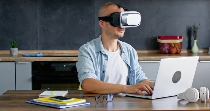 A bald man in 3D virtual reality glasses sits in front of a laptop. The work of the future in virtual reality.