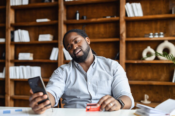 Displeased African-American man lholding credit card and smartphone, reading rejected message, guy has bad balance, bankruptcy, debt