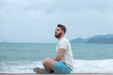 Fototapeta na wymiar Young calm sad serious man is sitting on embankment near sea, ocean on beach, thinking, meditate on natural background 