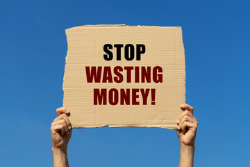 Stop wasting money text on box paper held by 2 hands with isolated blue sky background. This message board can be used as business concept about stop wasting money.