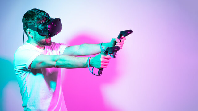 Bearded man in VR glasses plays in simulation figth and using controllers in in multicolored neon lights colorful background. Concept interactive online game in a virtual world. Copy space, banner