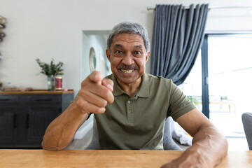 Portrait of cheerful biracial senior man pointing at camera while talking over video call at table