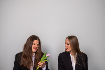 Couple in suits. Disappointed man with long, brown hair holding a tulip. Woman looking at a tulip in man's hand.