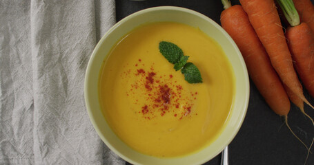 Close up of white bowl of carrot soup with bread, napkin and carrots on dark background