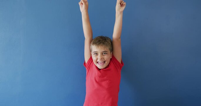 Portrait of happy caucasian boy raising hands and smiling on blue background, slow motion