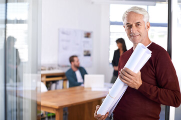 Design minds at work. Portrait of a mature male architect holding blueprints with his colleagues working in the background.