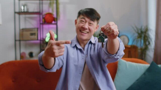 Asian man real estate agent lifting hand showing the keys of new home house apartment, buying or renting property, mortgage loan at modern home apartment indoors. Chinese guy in room sitting on sofa
