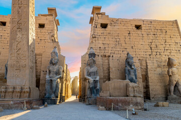 The entrance to the ancient Egyptian Luxor Temple with statues of Rameses II and the pylon obelisk as the sun turns to colors near sunset in Luxor, Egypt.