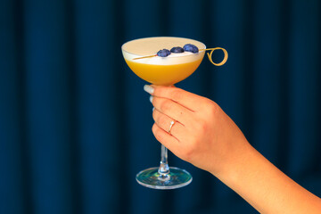 A cocktail in a glass with a long stem in a woman's hand on a blurred background of a blue soft...