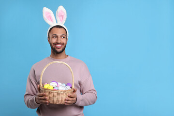 Happy African American man in bunny ears headband holding wicker basket with Easter eggs on light...