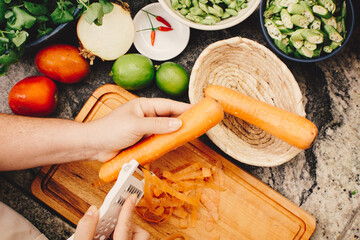 Top-down photo of female hand peeling a carrot with a manual peeler on a wooden cutting board...