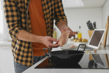 Man putting chicken fillet into frying pan while watching online cooking course via laptop in kitchen, closeup